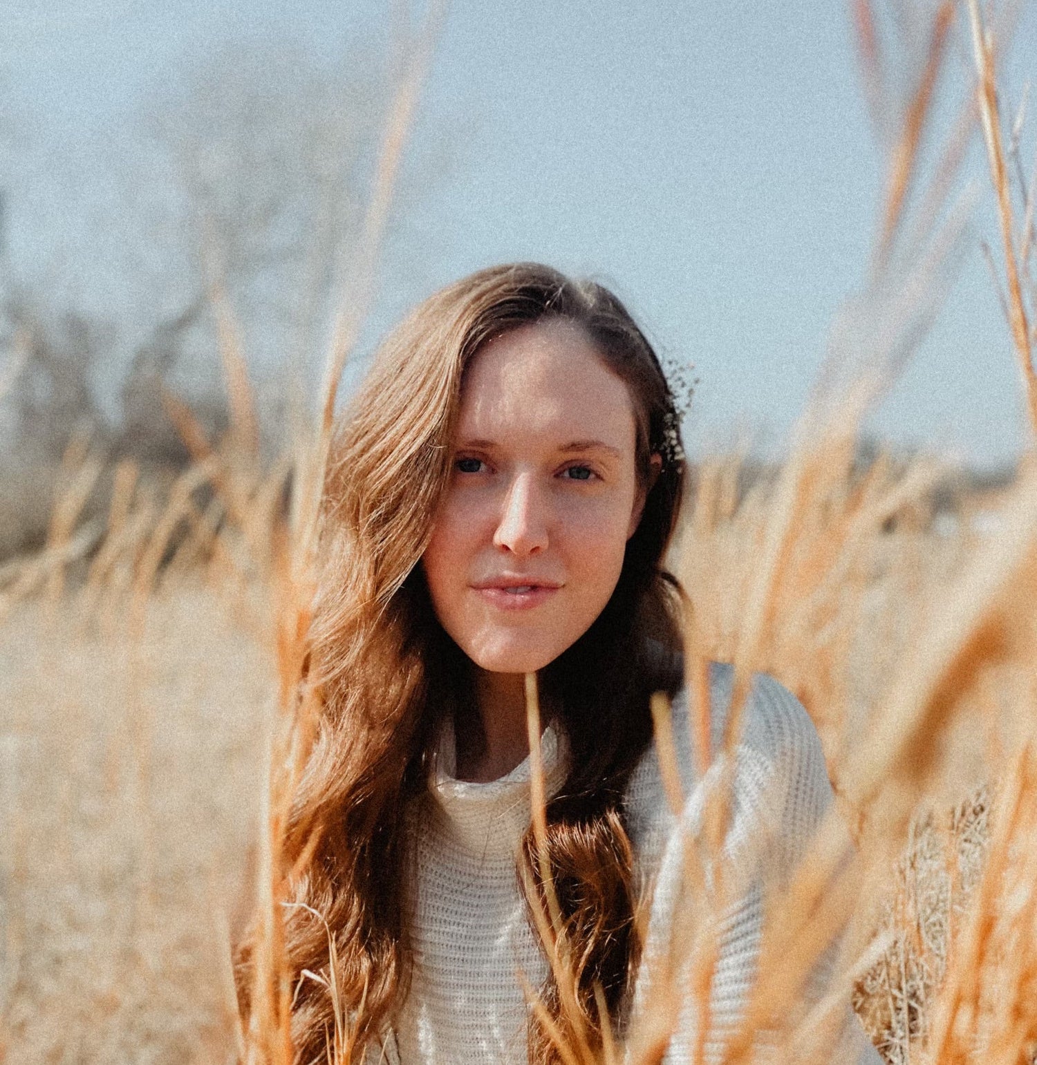 Woman with long brown hair sitting among tall grasses with a clear blue sky in the background